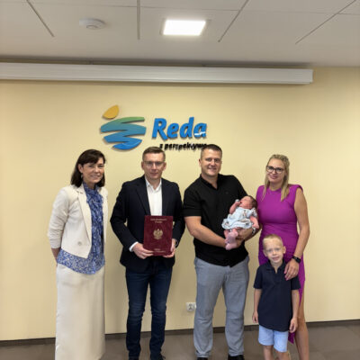 Na zdjęciu widzimy pięć osób stojących na tle żółtej ściany z logotypem miasta Reda oraz hasłem „Reda z perspektywą”. Po lewej stronie stoi kobieta w jasnym garniturze i niebieskiej koszuli. Obok niej stoi burmistrz w granatowej marynarce i jeansach, trzymający czerwoną teczkę z godłem Rzeczypospolitej Polskiej. Obok niego – uśmiechnięty mężczyzna w czarnej koszulce i jasnych spodniach, trzymający na rękach noworodka w jasnej sukience. Po prawej stronie znajduje się kobieta w różowej sukience oraz chłopiec, który stoi przodem do aparatu – to starszy brat Helenki. Wszyscy patrzą w stronę obiektywu i uśmiechają się.